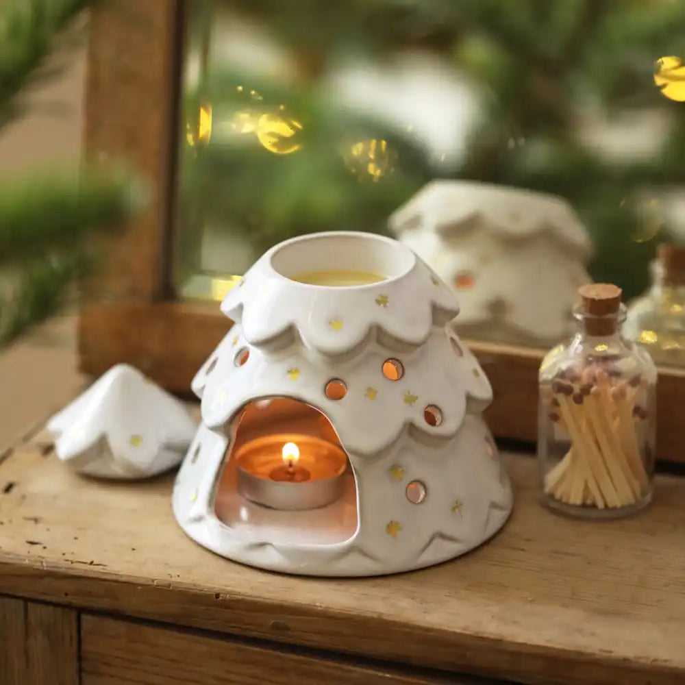 A white ceramic winter tree-shaped was burner with star details, shown lit with a tea light candle inside, set on a wooden surface with a glass bottle of matches nearby.