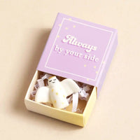 Small white figurine in a box with 'Always by your side' text on a beige background