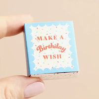 A hand holding a small square gift box with a light blue border, white center, and the words “MAKE A Birthday WISH” in coral script with multicolored confetti. The background is a soft beige.