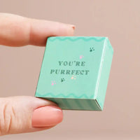 Small green box with 'YOU'RE PURRFECT' text held by a hand against a beige background