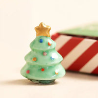 Small green Christmas tree figurine with a gold star on top, set against a blurred red and white striped background.