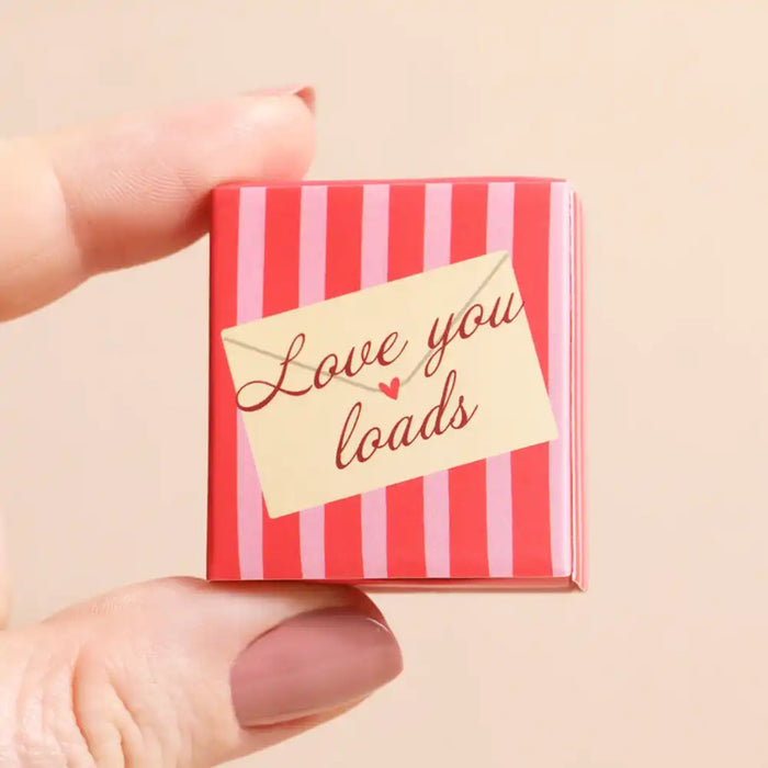 A hand holding a tiny red and pink striped square box with a cream envelope-style label that reads 'Love you loads' in red script, against a soft beige background.