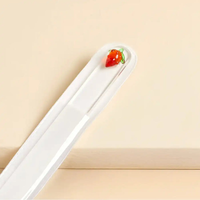Glass nail file with glass strawberry charm decoration on a beige background.