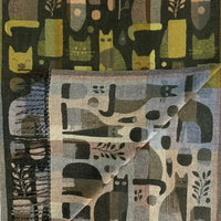 A folded woven textile featuring an abstract cat-themed pattern in muted olive, mustard, gray, beige, and charcoal tones, with visible fringe on one edge.