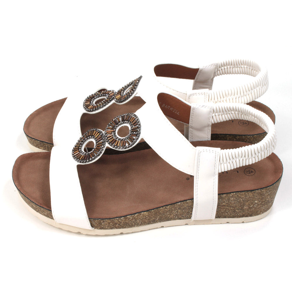White sandals with medium cork heels. Jewelled circular decoration on the straps. Elasticated heel straps. Side view.