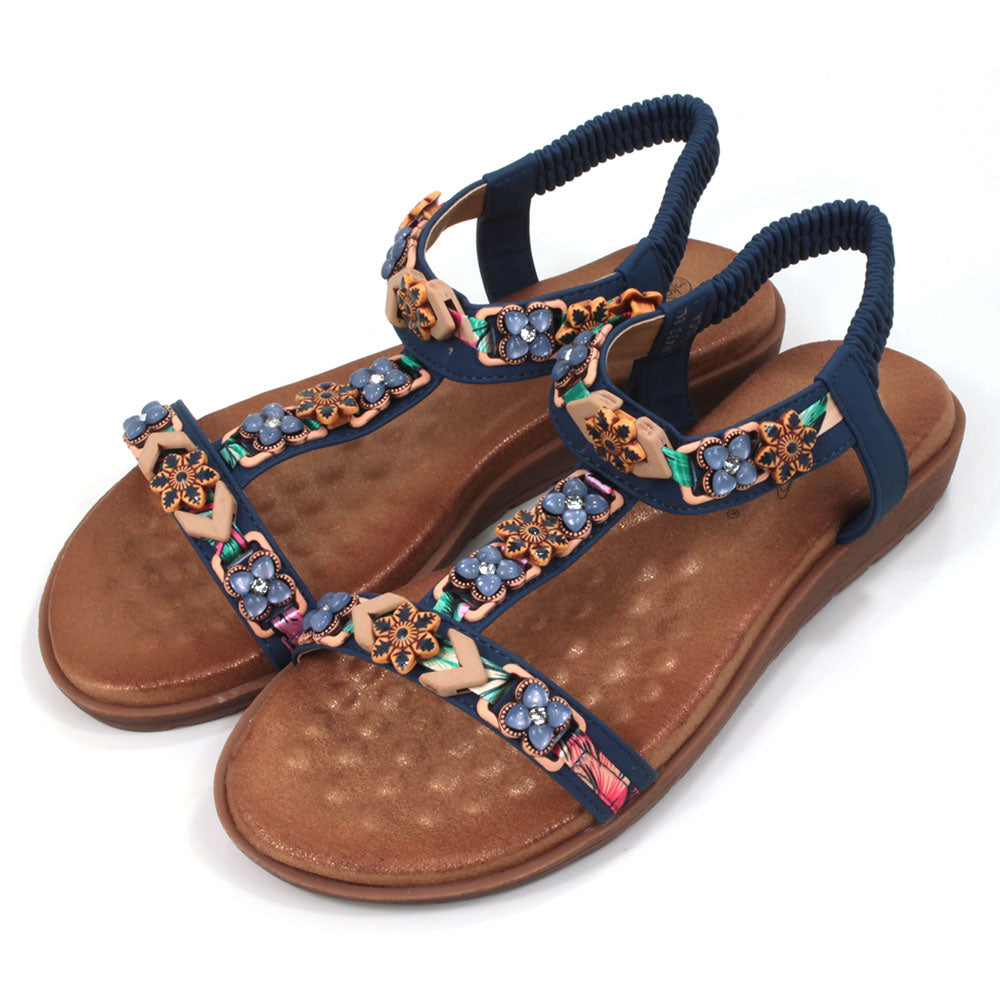 Flat sandals  with padded tan coloured footbeds. Blue straps including elasticated ankle straps.  Decorated with a large assortment of beads and other decorative elements. Angled view.