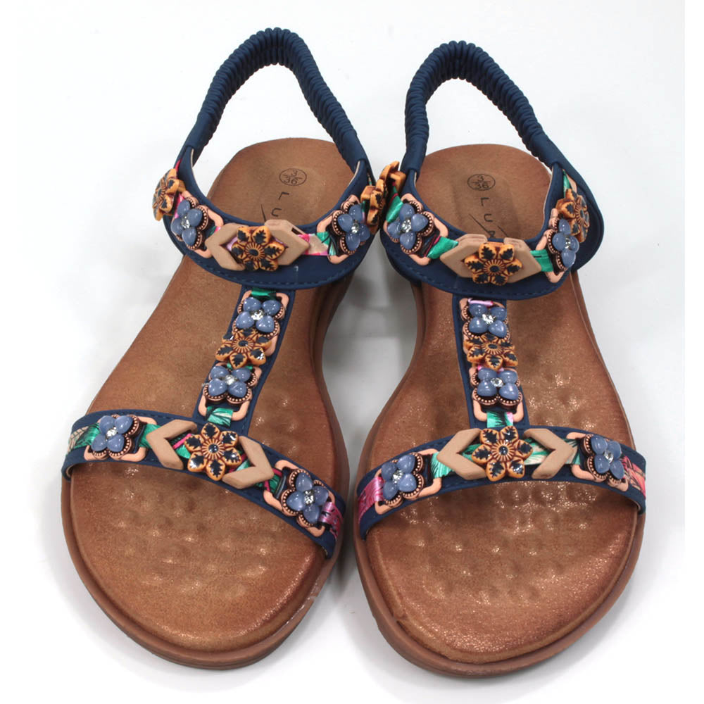 Flat sandals  with padded tan coloured footbeds. Blue straps including elasticated ankle straps.  Decorated with a large assortment of beads and other decorative elements. Front view.