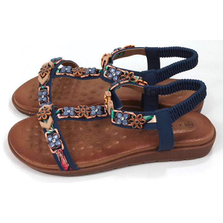 Flat sandals  with padded tan coloured footbeds. Blue straps including elasticated ankle straps.  Decorated with a large assortment of beads and other decorative elements. Side view.