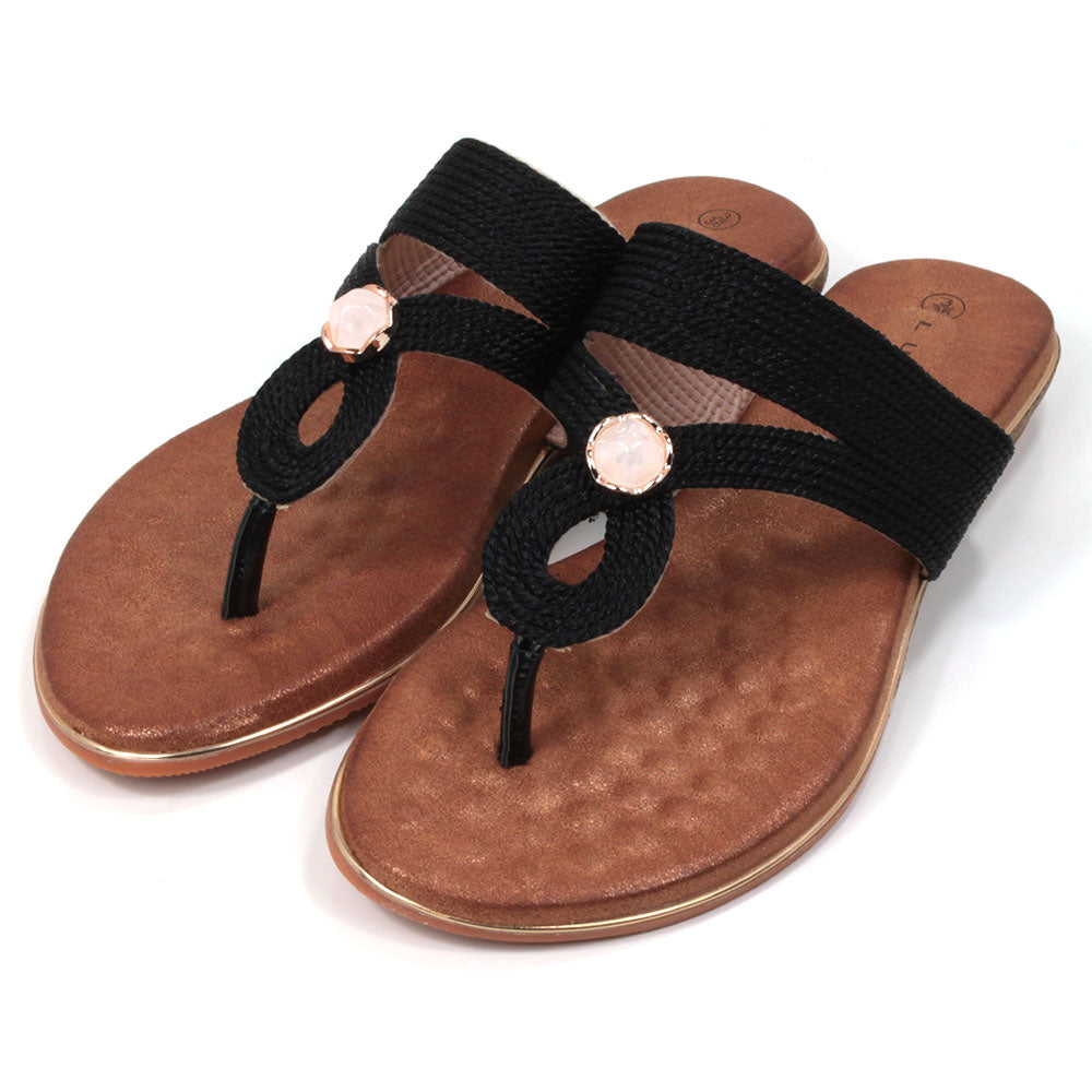 Flat toe post sandals in black with tan coloured textured footbed. Straps in textured fabric and featuring a gemstone centred over the foot. Angled view.