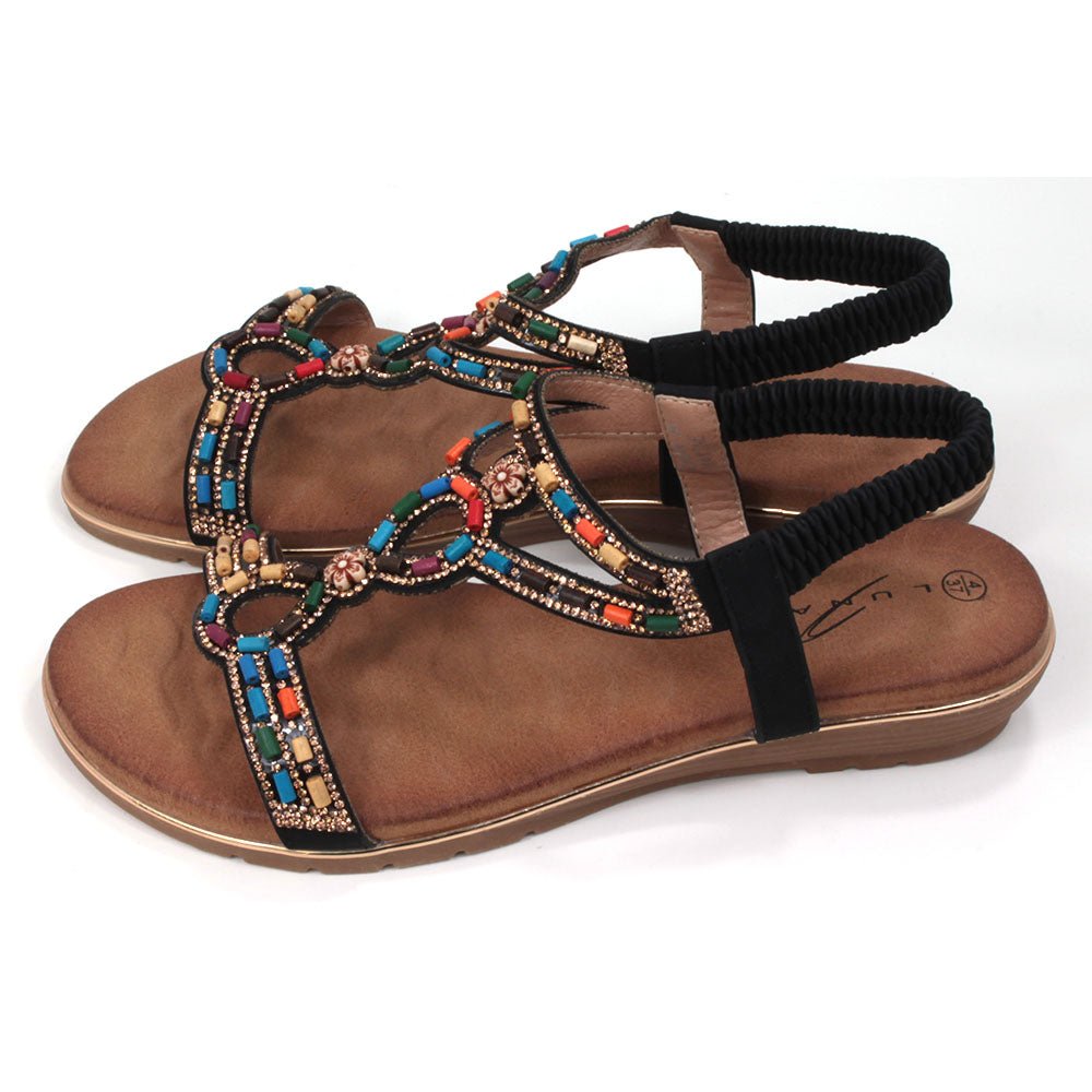 Strappy sandals with elasticated ankle strap in black. Decorated with colourful beads. Tan coloured footbeds. Flats. Side view.