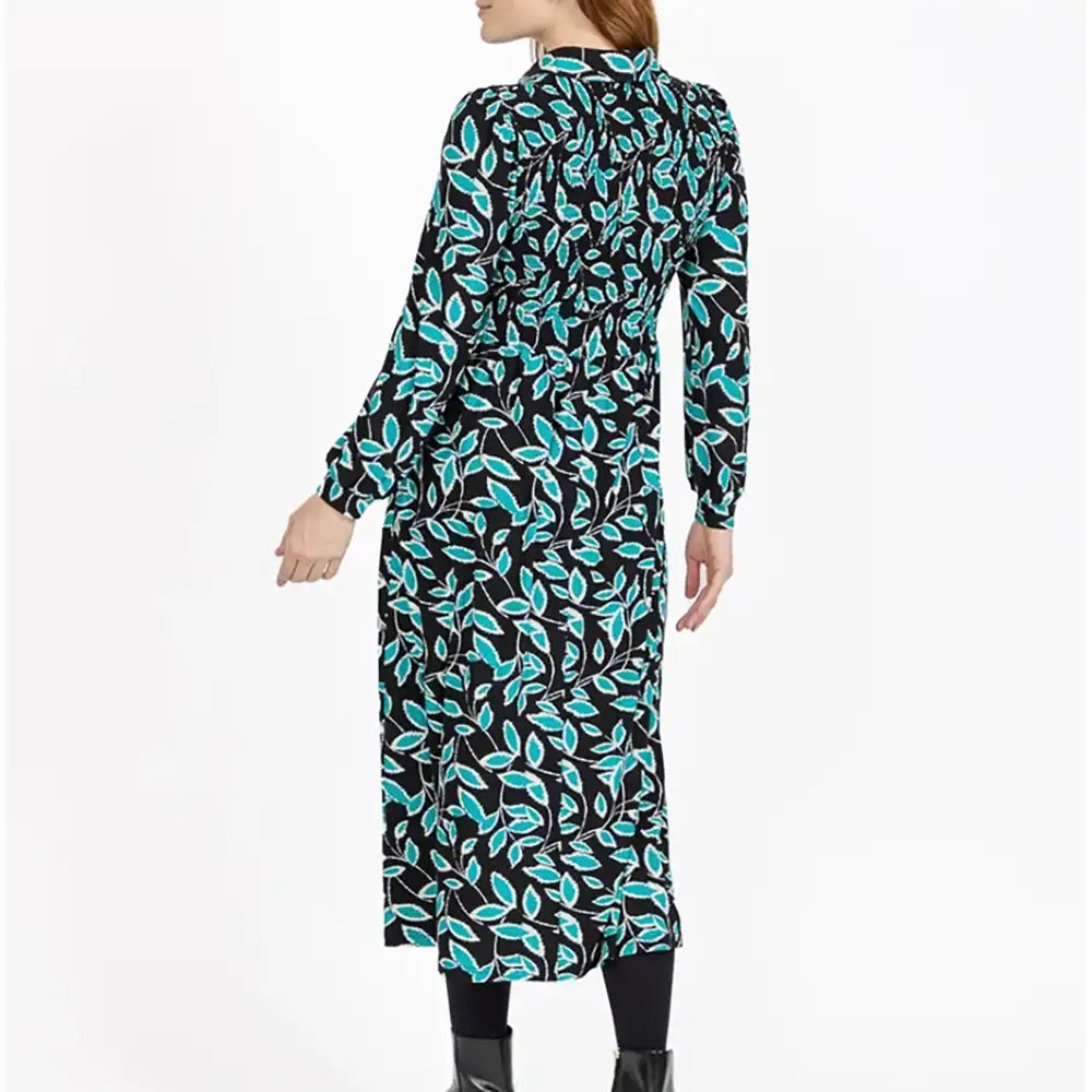 A woman modeling a long-sleeve midi dress with a black background and a vibrant turquoise and white leaf pattern, shown from a rear three-quarter view against a plain white studio background.