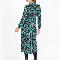 A woman modeling a long-sleeve midi dress with a black background and a vibrant turquoise and white leaf pattern, shown from a rear three-quarter view against a plain white studio background.