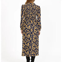 A woman viewed from the back wearing a long-sleeve midi dress with a navy blue base and an all-over mustard yellow leaf pattern, paired with black pants, against a plain white studio background.