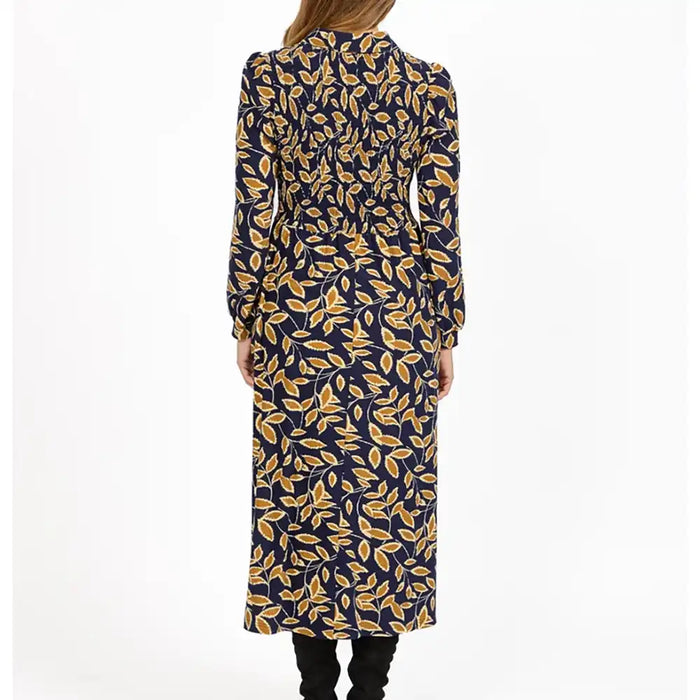 A woman viewed from the back wearing a long-sleeve midi dress with a navy blue base and an all-over mustard yellow leaf pattern, paired with black pants, against a plain white studio background.