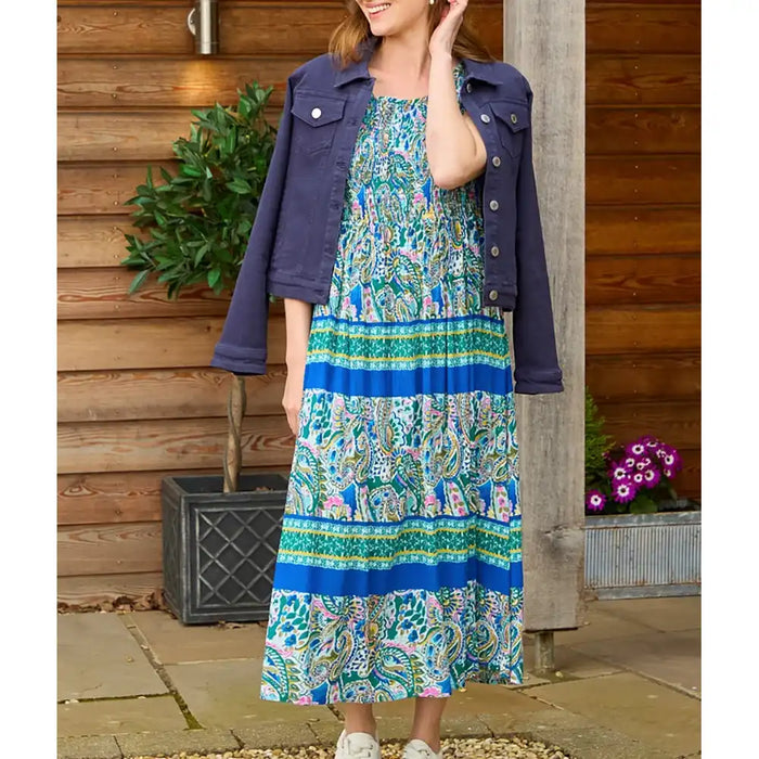 A woman wearing a blue and green paisley maxi dress with a dark blue denim jacket draped over her shoulders, standing in front of a wooden wall.