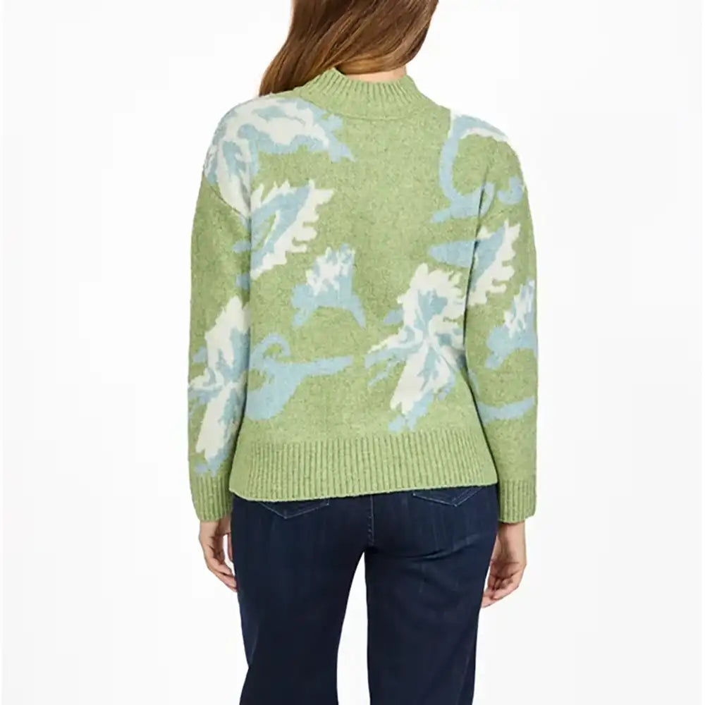 A woman viewed from the back wearing a light green knit sweater with a large white and light blue abstract floral pattern, paired with dark blue jeans, against a plain white background.