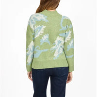 A woman viewed from the back wearing a light green knit sweater with a large white and light blue abstract floral pattern, paired with dark blue jeans, against a plain white background.