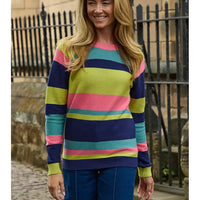 A woman wearing a colorful striped knit sweater with navy, pink, teal, and lime green horizontal stripes, paired with blue jeans, standing outdoors in front of a black metal fence.