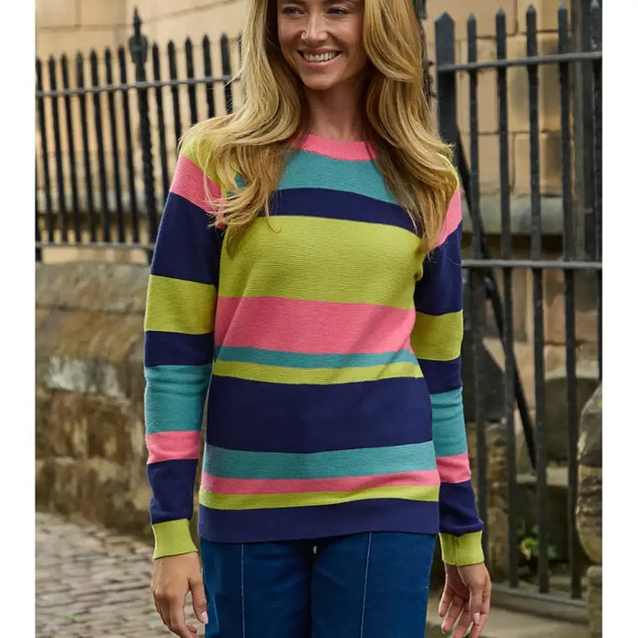 A woman wearing a colorful striped knit sweater with navy, pink, teal, and lime green horizontal stripes, paired with blue jeans, standing outdoors in front of a black metal fence.