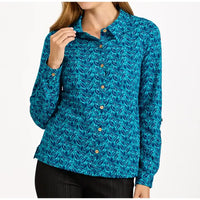 A woman wearing a long-sleeve button-up shirt with a blue and teal abstract pattern, paired with black pants, against a plain white background.