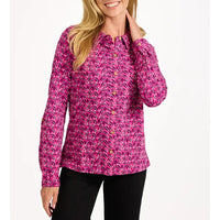 A woman wearing a long-sleeve button-up blouse with a vibrant magenta and pink tropical floral pattern, paired with black pants, against a plain white background.