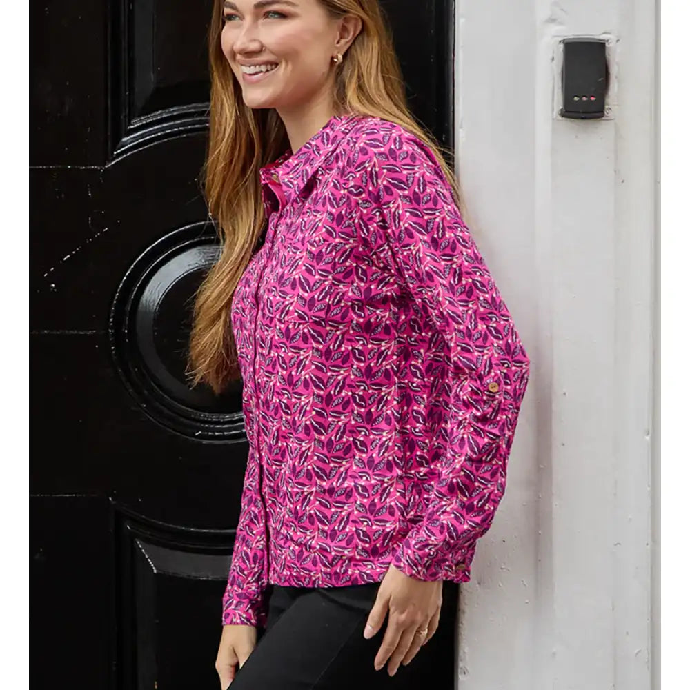 A woman wearing a pink long-sleeve button-up shirt with a white and yellow abstract pattern, standing next to a black door and a white wall.