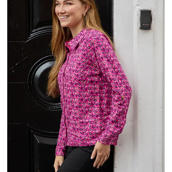 A woman wearing a pink long-sleeve button-up shirt with a white and yellow abstract pattern, standing next to a black door and a white wall.