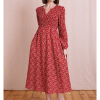 A woman wearing a red long-sleeve midi dress with a white abstract floral pattern, standing against a beige fabric backdrop and wooden floor.