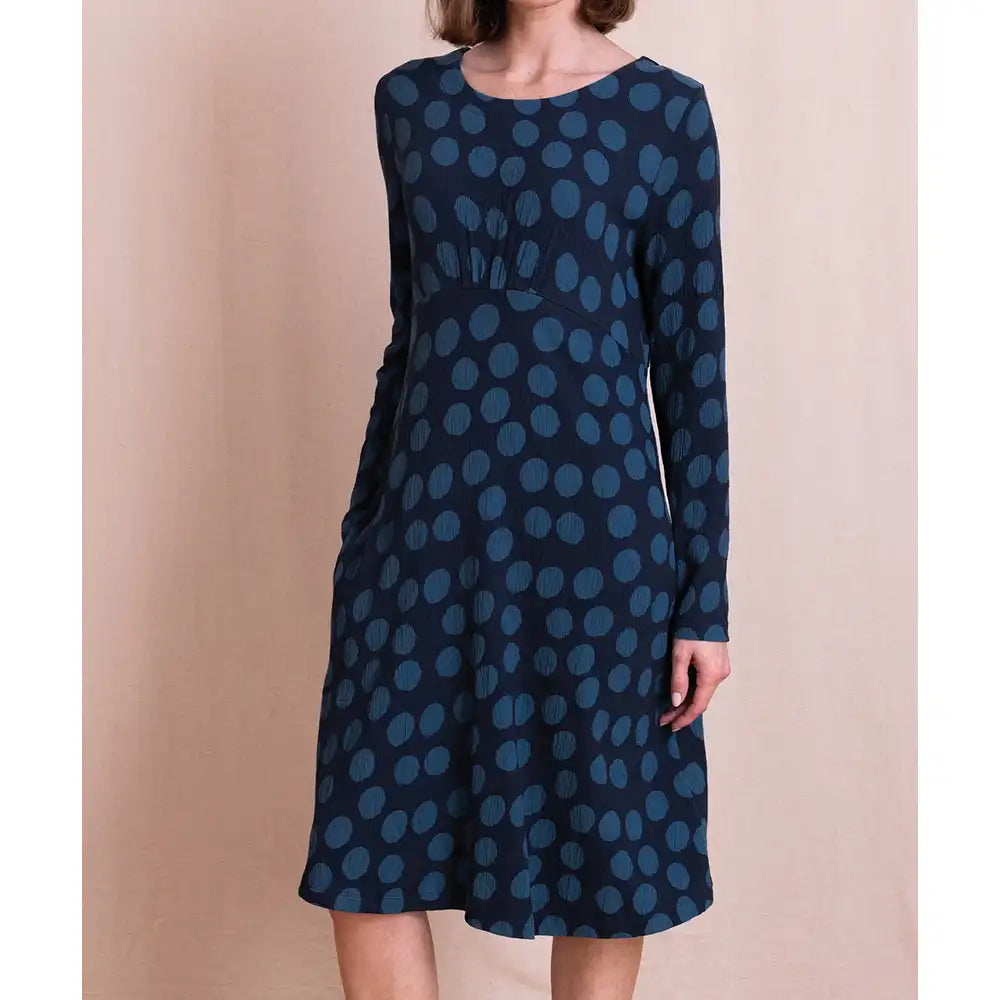 A woman wearing a long-sleeve knee-length dress with a dark navy base and a lighter blue polka dot pattern, standing against a solid beige backdrop.