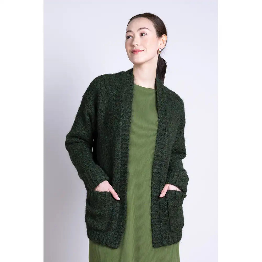 Woman wearing a chunky dark green cardigan with front patch pockets over a green dress on a white background