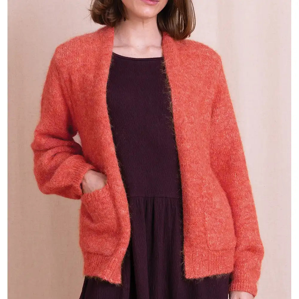 A woman wearing a fuzzy, textured coral-orange cardigan with side pockets over a dark plum dress, set against a solid beige background.