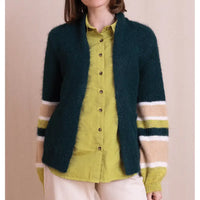 A person wearing a fuzzy dark teal cardigan over a yellow-green button-up shirt and cream pants, photographed against a plain beige studio background.