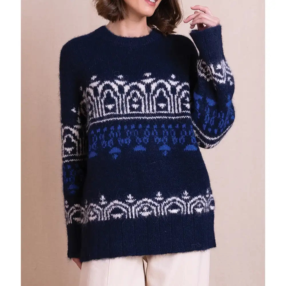 A woman wearing a fuzzy navy blue knit sweater with a white and light blue Nordic/Fair Isle pattern across the chest, sleeves, and hem, paired with light beige pants against a solid beige background.