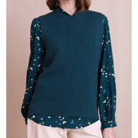 A woman wearing a dark teal ribbed knit mock-neck sweater vest over a matching dark teal blouse with a small multicolor butterfly print, paired with light beige pants.