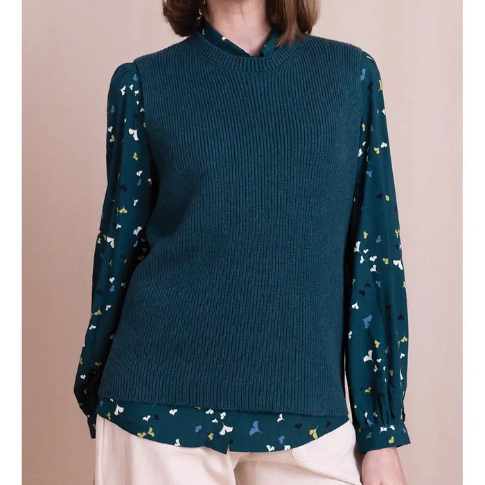 A woman wearing a dark teal ribbed knit mock-neck sweater vest over a matching dark teal blouse with a small multicolor butterfly print, paired with light beige pants.