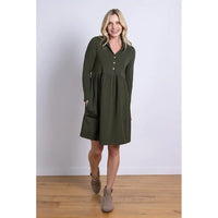 Woman wearing a green tunic style dress with hands in pockets standing on a wooden floor with a white wall background