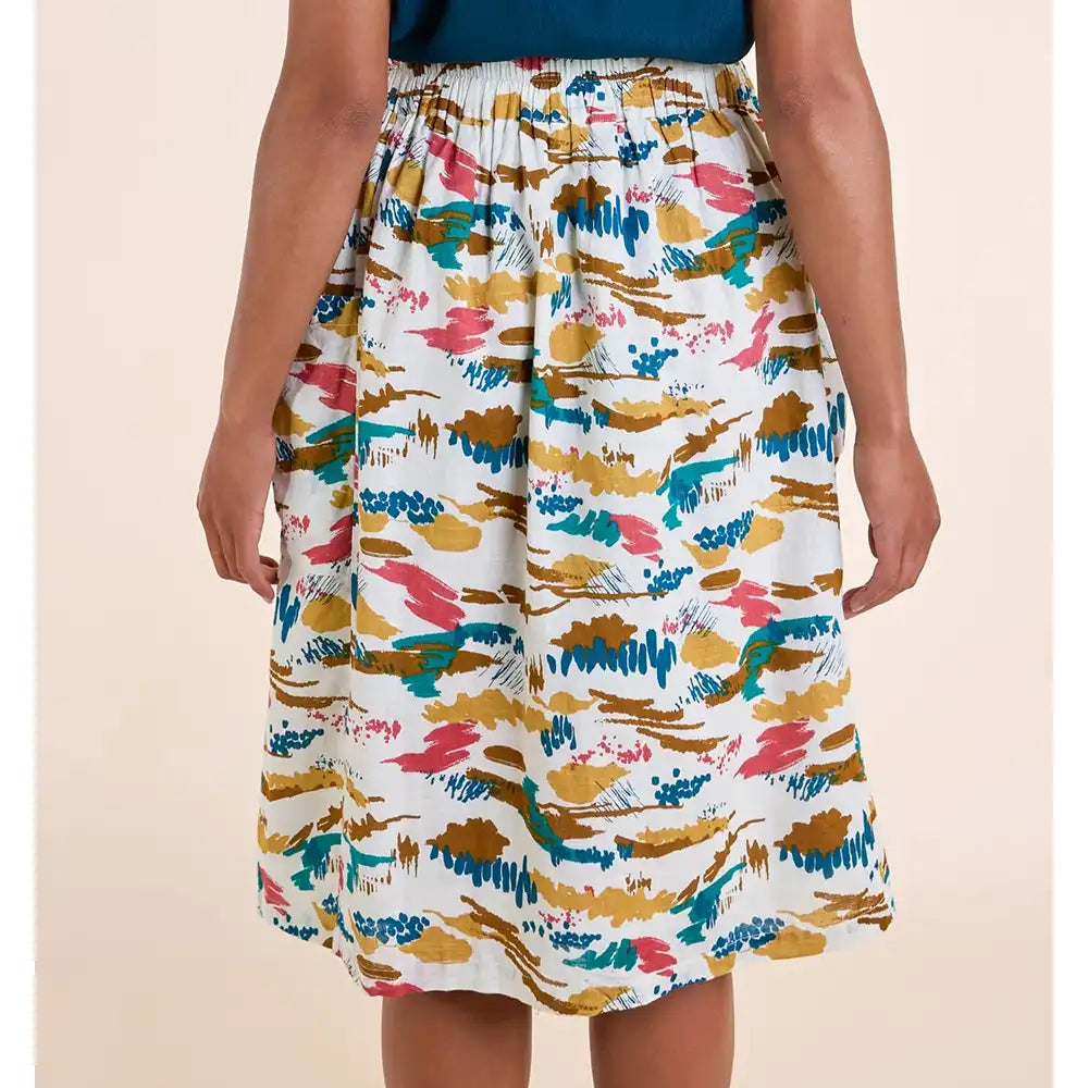 A model wearing a white A-line midi skirt with a colorful abstract brushstroke pattern in teal, mustard yellow, pink, and brown, paired with a dark blue top against a solid beige background.