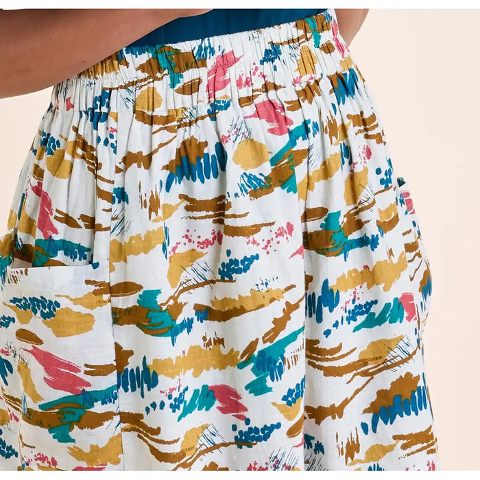 Close-up of a person wearing high-waisted, elastic-waist shorts with a colorful abstract brushstroke print on a white background. The print features gold, teal, pink, and blue strokes, and a side pocket is visible on the left leg.