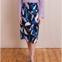 A model wearing a navy blue knee-length pencil skirt with an abstract brushstroke pattern in blue, purple, and white, paired with a lavender sweater and silver heels, standing on a wooden floor against a beige backdrop.