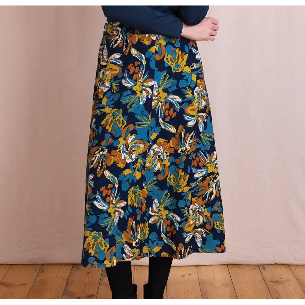 A person wearing a navy midi skirt with a bold yellow, orange, and white floral print, paired with a dark navy top and black tights, standing against a beige fabric backdrop and wooden floor.