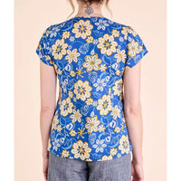 A woman shown from the back wearing a short-sleeve blue top with a yellow and white floral pattern, paired with gray pants, against a solid beige background.