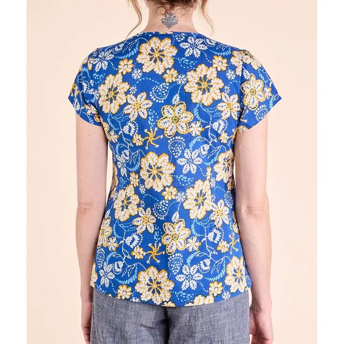 A woman shown from the back wearing a short-sleeve blue top with a yellow and white floral pattern, paired with gray pants, against a solid beige background.