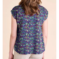 A woman shown from the back wearing a short-sleeve top with a dense multicolor vertical brushstroke pattern in purple, blue, green, and yellow on a dark base, paired with light beige pants, set against a solid pale peach background.