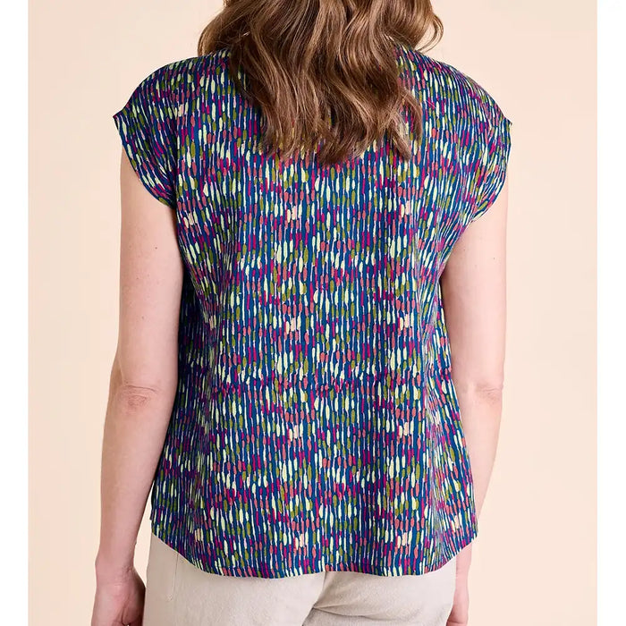 A woman shown from the back wearing a short-sleeve top with a dense multicolor vertical brushstroke pattern in purple, blue, green, and yellow on a dark base, paired with light beige pants, set against a solid pale peach background.