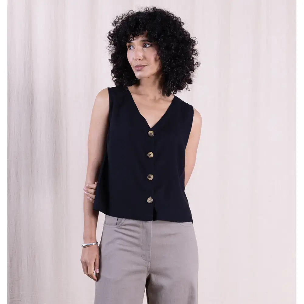 Woman wearing a black sleeveless top with gold buttons against a light background