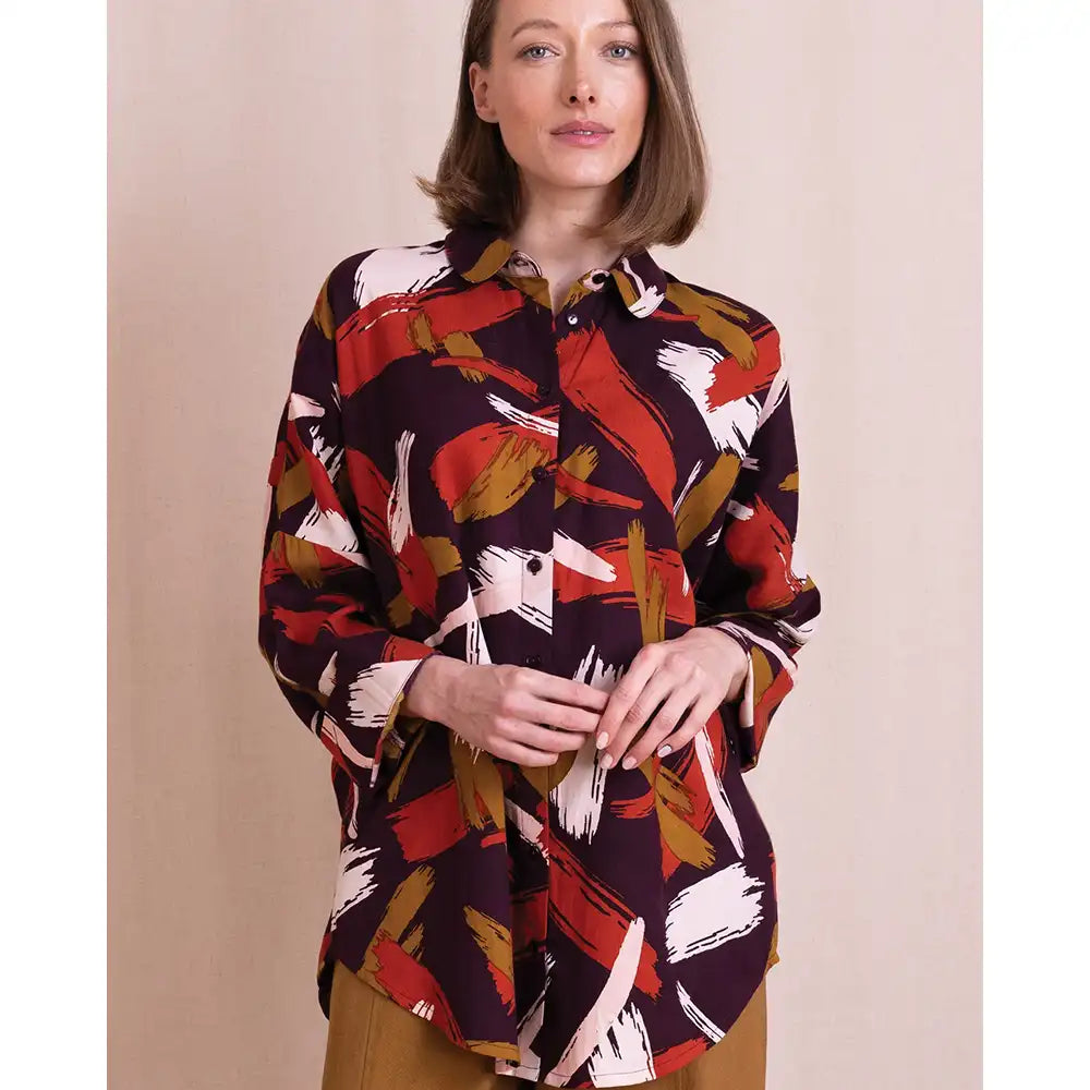 A woman wearing a long-sleeve button-up shirt with an abstract brushstroke pattern in red, white, and brown on a dark background, paired with brown pants, set against a plain beige studio backdrop.