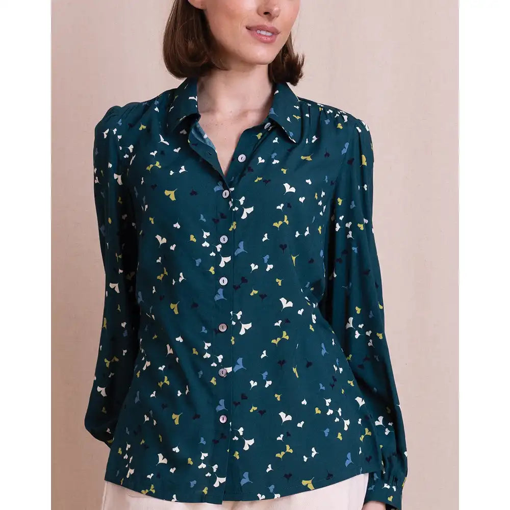 A woman wearing a dark teal long-sleeve button-up blouse with a small multicolor bird print, paired with light beige bottoms, set against a solid beige background.