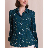 A woman wearing a dark teal long-sleeve button-up blouse with a small multicolor bird print, paired with light beige bottoms, set against a solid beige background.