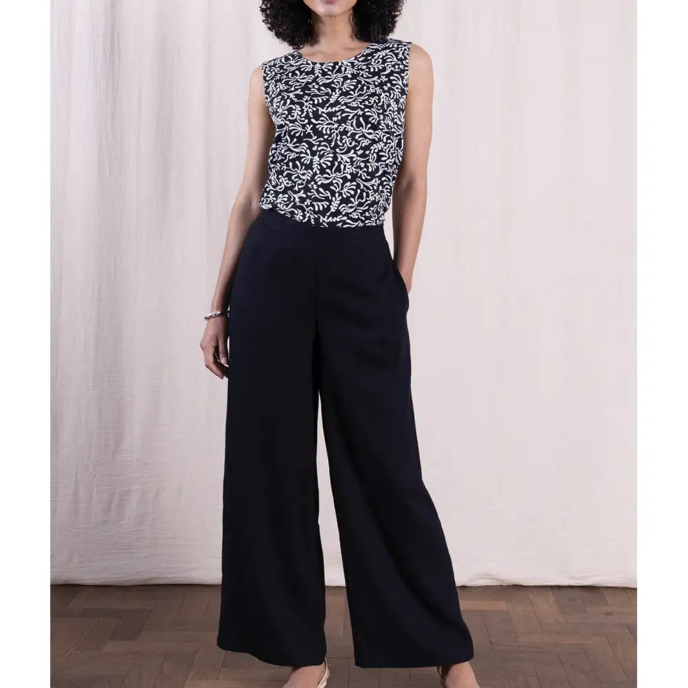 Person wearing a sleeveless patterned top and black wide-leg pants against a white curtain background