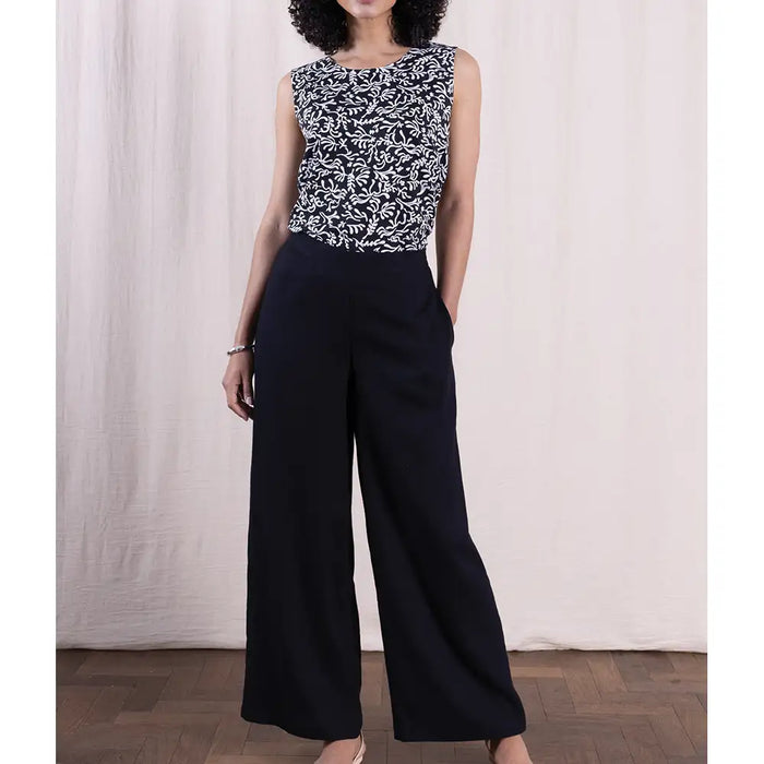 Person wearing a sleeveless patterned top and black wide-leg pants against a white curtain background